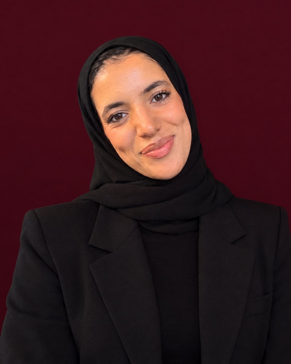 Portrait of Maryam Ibaaichou wearing a black hijab and blazer against a deep burgundy background